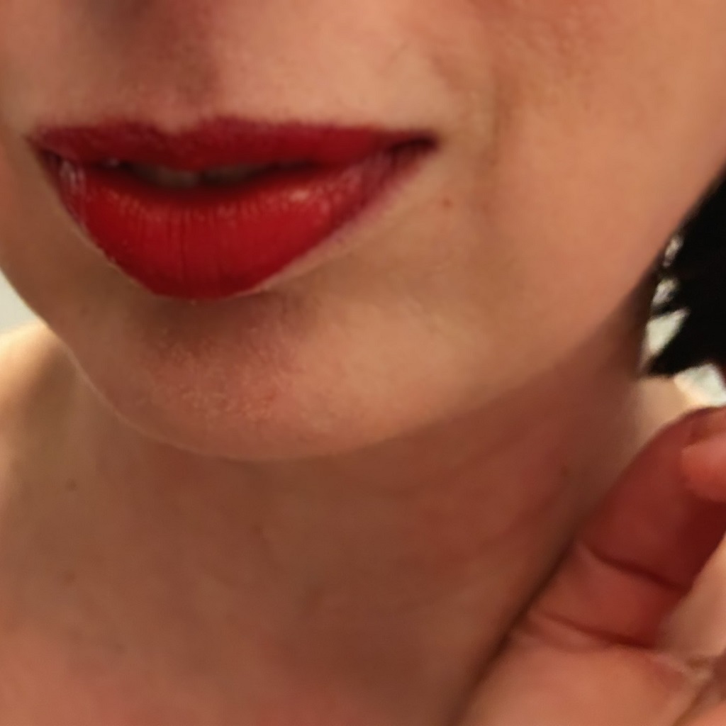Close-up of my lips. They're painted red and shiny. It looks like the person I'm observing just did something mildly amusing and definitely intriguing. My hand is palm up beside my chin slightly curled, index finger and thumb touching. Chin length dark hair is visible. Everything is ever so slightly out of focus. The colours are warm.
